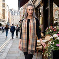 Elegant model wearing Camel Thomson Pashmina Scarf with tartan pattern outdoors in golden hour, showcasing its soft, silky texture and stylish design.