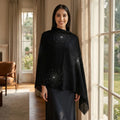Model wearing embroidered pashmina shawl over dress by large window in luxury home, showcasing delicate embroidery and drape.